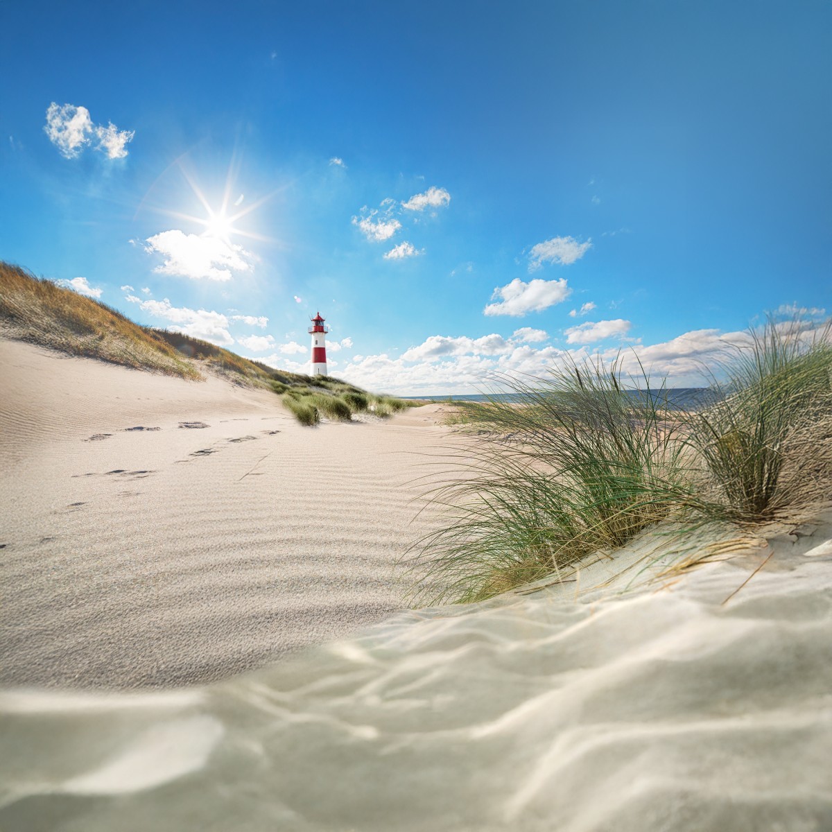Ein Leuchtturm steht auf einer Düne am Strand unter einem blauen Himmel mit Sonne und Wolken Fußspuren führen durch den Sand, im Vordergrund wachsen Gräser am Strand.