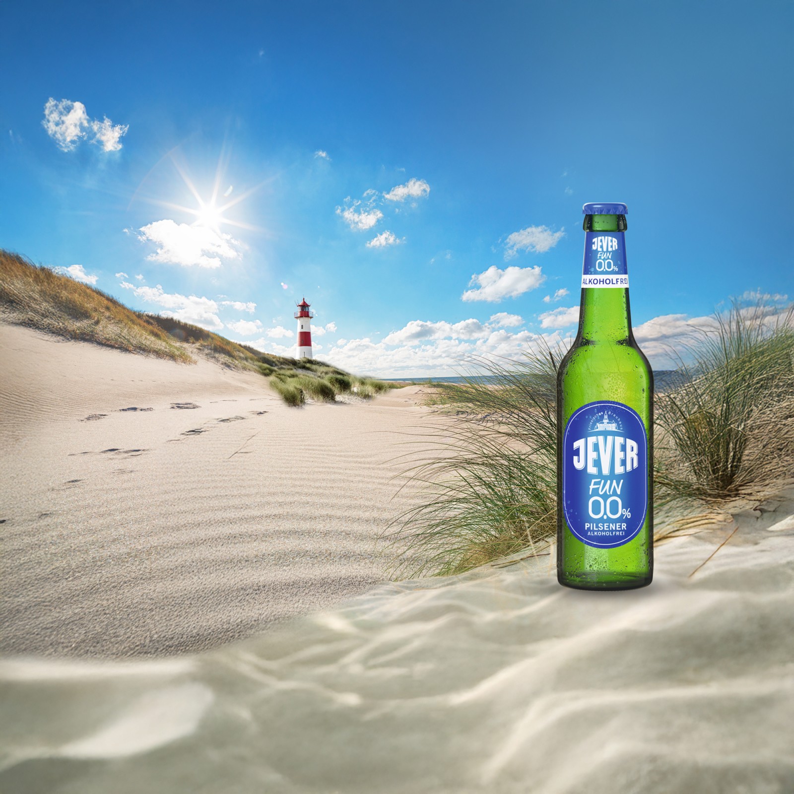 Ein Leuchtturm steht auf einer Düne am Strand unter einem blauen Himmel mit Sonne, im Vordergrund steht ein Flasche Jever Fun null komma null.