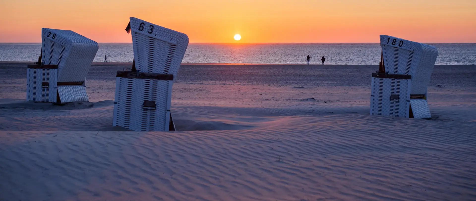 Drei Strandkörbe stehen auf sandigem Strand mit Sonnenuntergang im Hintergrund. Zwei Personen spazieren am Meer.