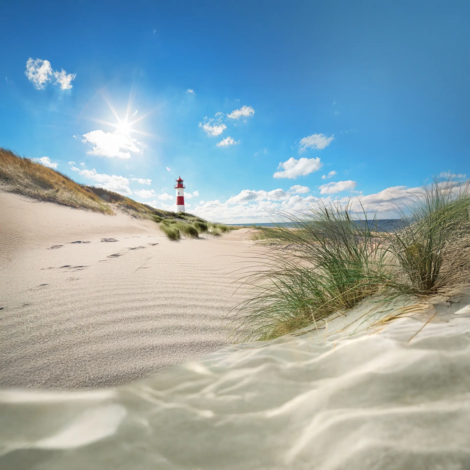 Ein Leuchtturm steht auf einer Düne am Strand unter einem blauen Himmel mit Sonne und Wolken Fußspuren führen durch den Sand, im Vordergrund wachsen Gräser am Strand.