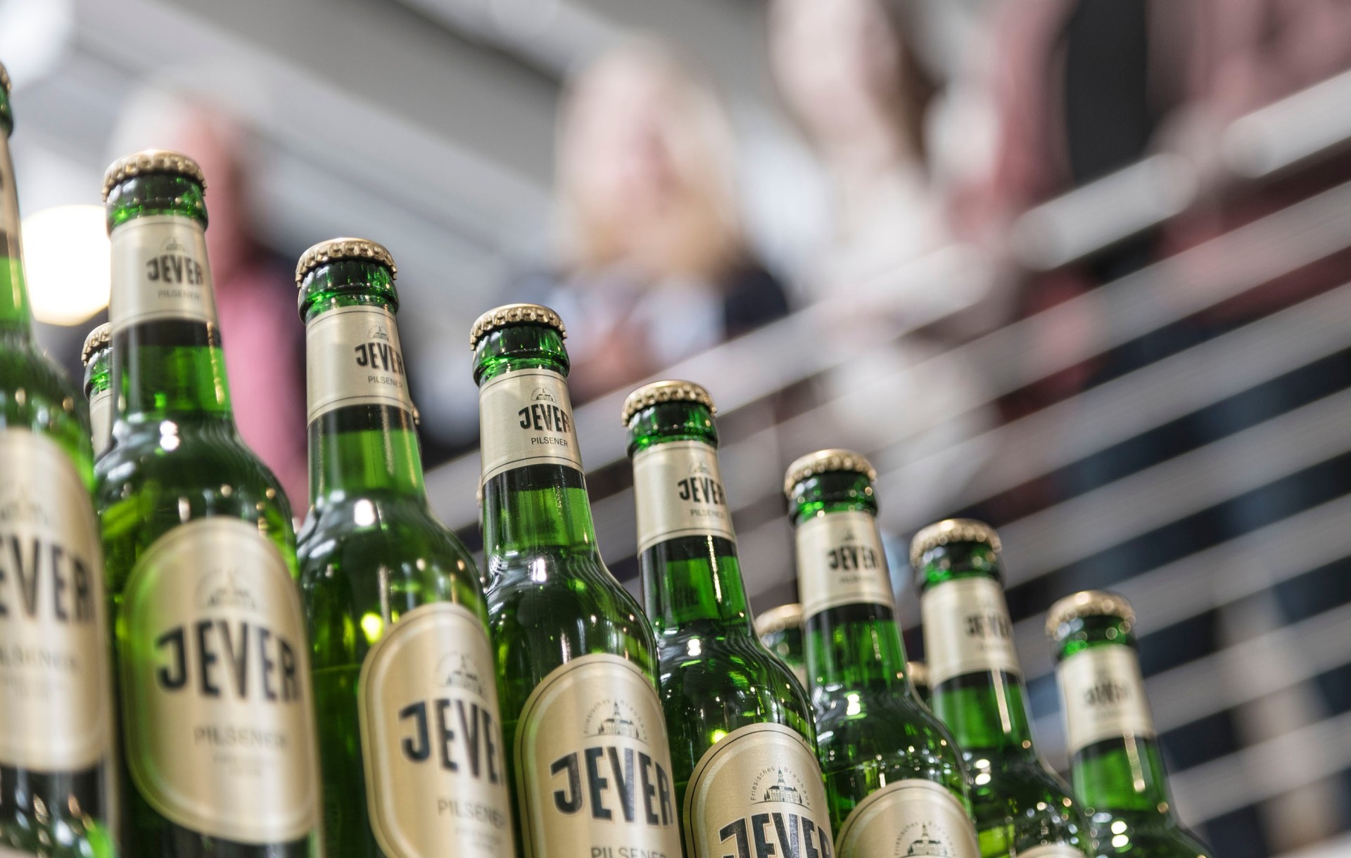 Grüne Bierflaschen stehen im Vordergrund, verschwommene Personen im Hintergrund.