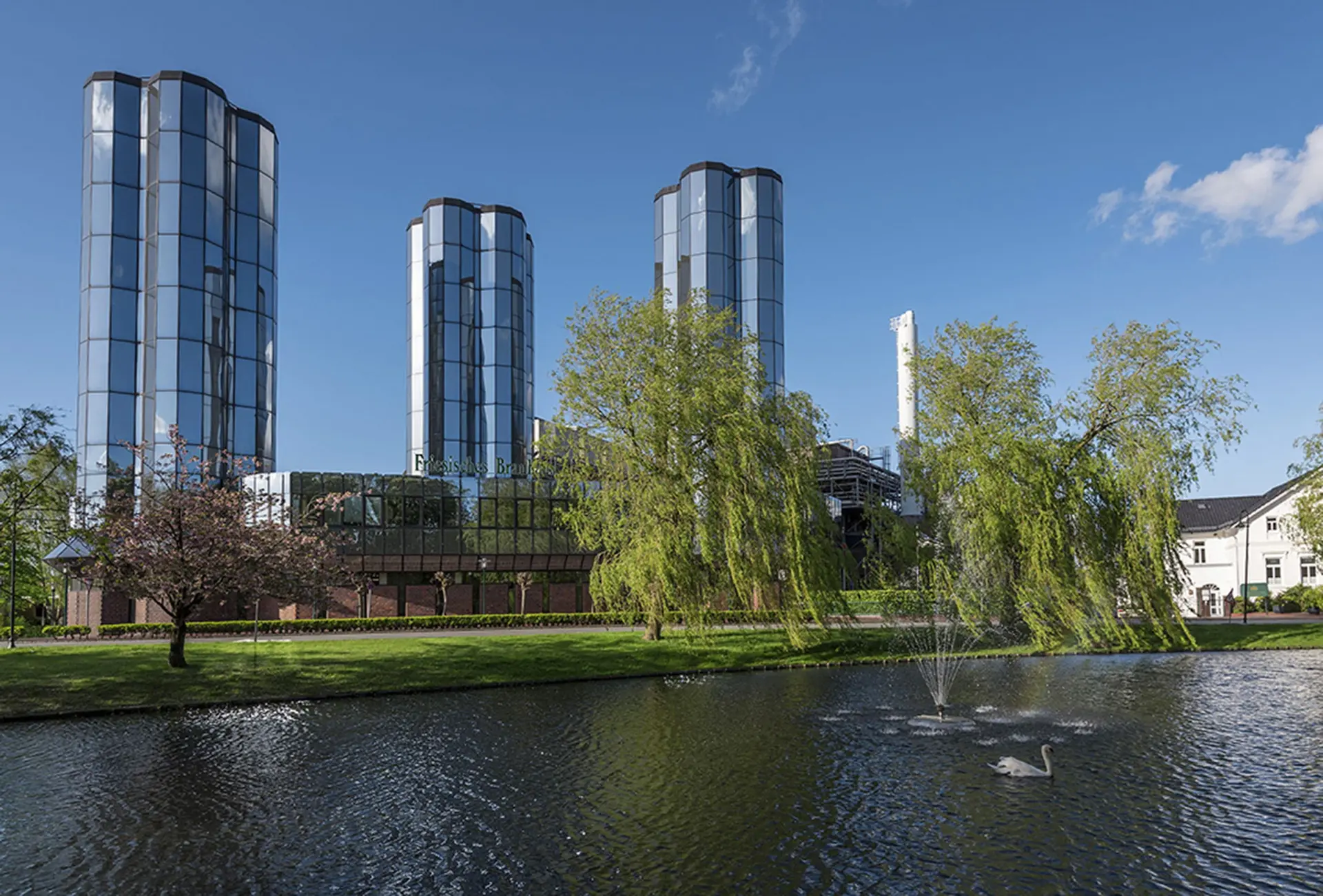Modernes Glasgebäude einer Brauerei hinter einem Teich mit Fontäne. Weidenbäume und Rasenfläche unter blauem Himmel.