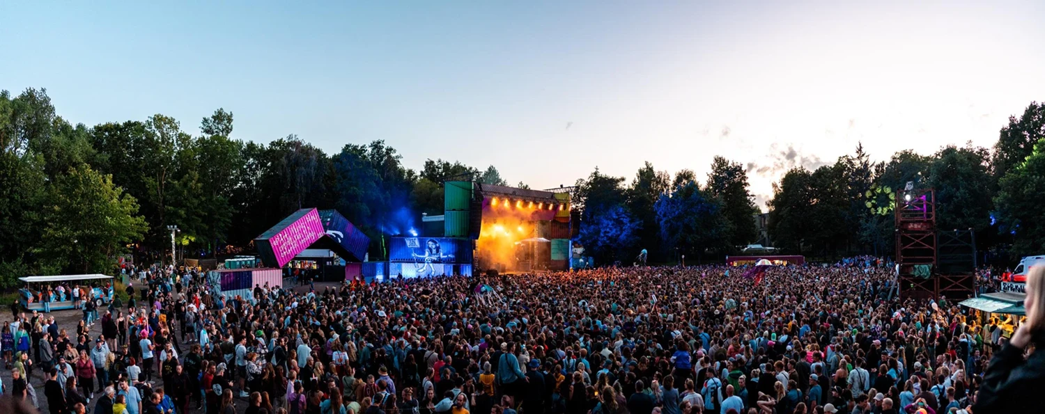 Großes Open-Air-Festival mit Menschenmenge vor einer Bühne. Im Hintergrund Bäume und blauer Himmel bei Sonnenuntergang.
