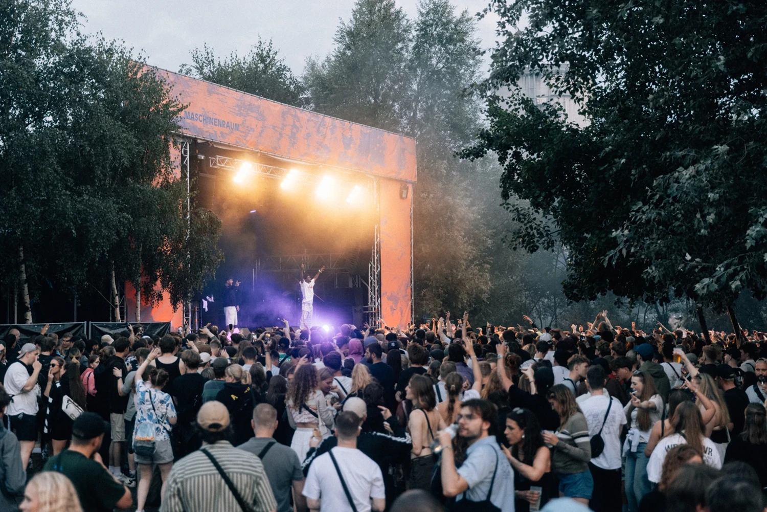 Menschenmenge bei Konzert vor einem beleuchteten orangefarbenen Bühnenaufbau.