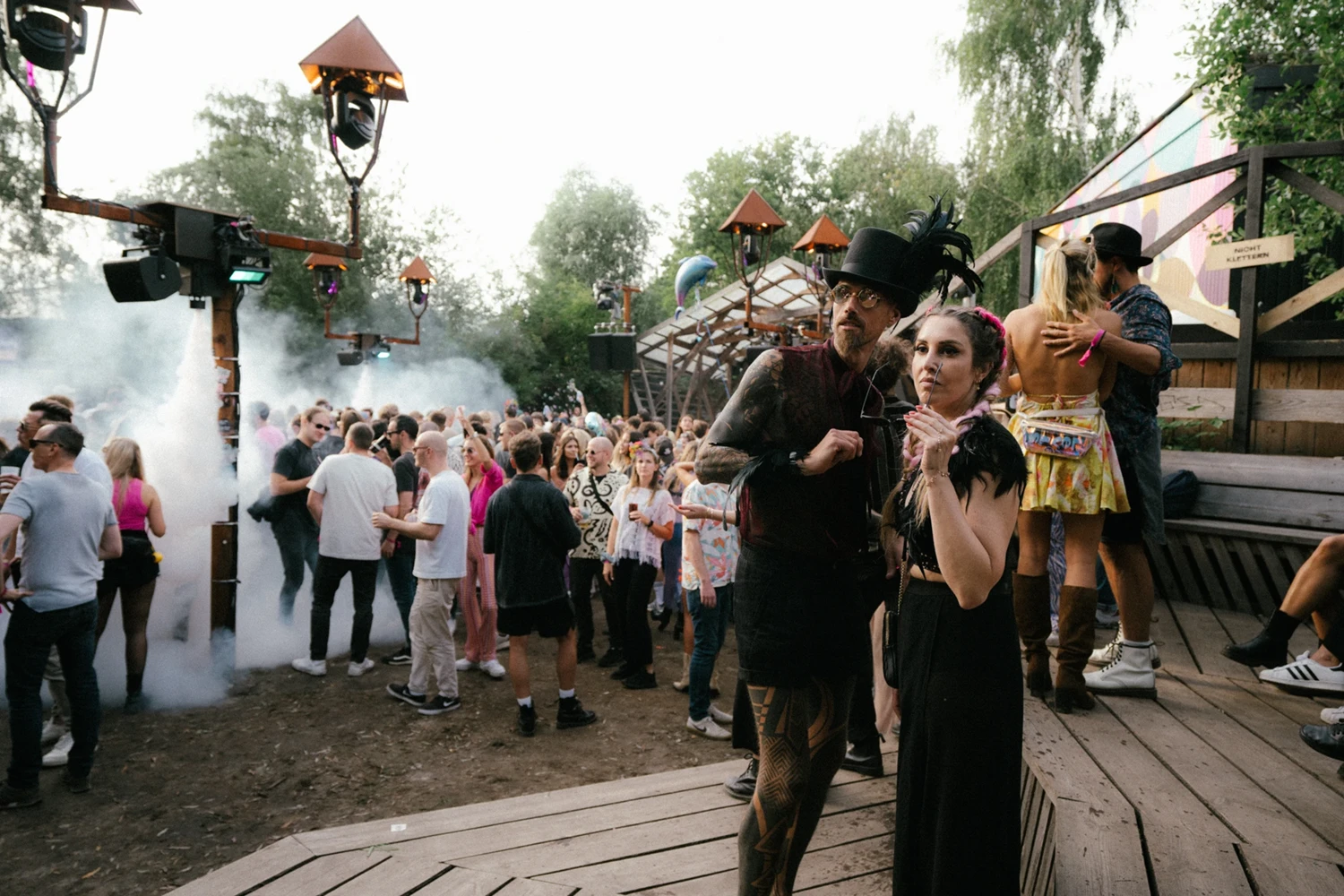 Festivalbesucher mit fantasievollen Outfits vor Tanzfläche mit Nebel. Im Hintergrund bewaldete Umgebung bei Tageslicht.