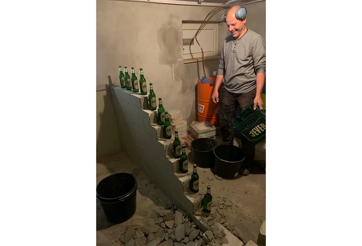 Eine gemauerte Treppe ist mit elf Jever Flaschen bestückt. Daneben steht ein lachender Mann am Arbeiten mit Gehörschutz.