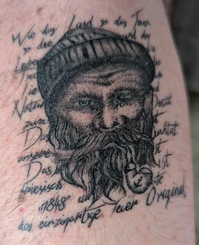 Ein Porträttattoo zeigt ein Gesicht mit Bart und Mütze auf Haut, umgeben von Text.