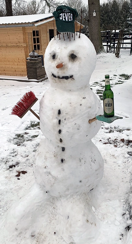 Ein Schneemann steht im Schnee mit einem grünen Hut und einer Flasche auf der Hand.