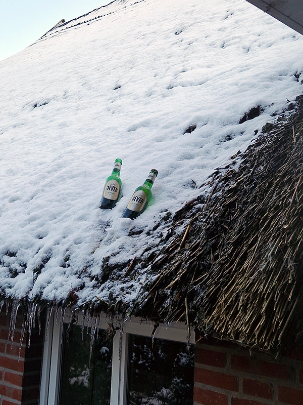Zwei leere Flaschen stehen auf einem schneebedeckten Strohdach über einem Fenster.