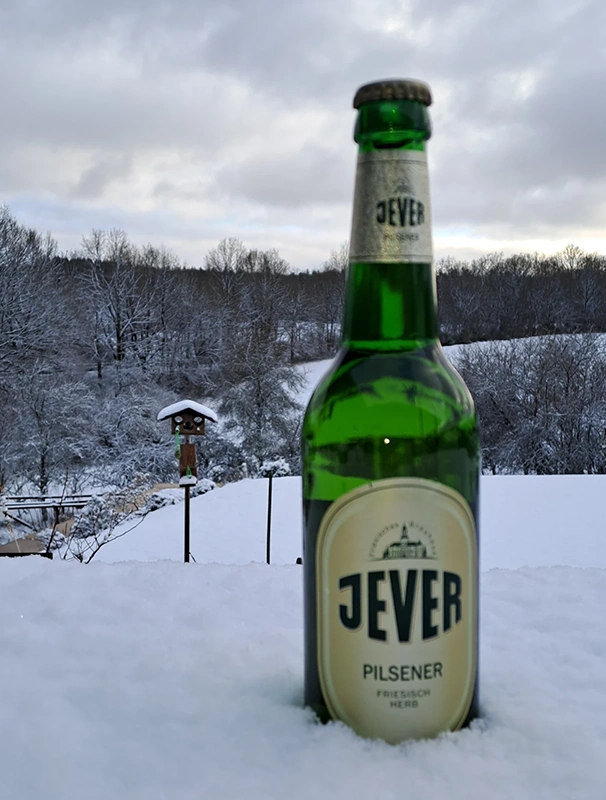 Eine Bierflasche steht im Vordergrund auf Schnee, dahinter Wald unter Schneedecke.