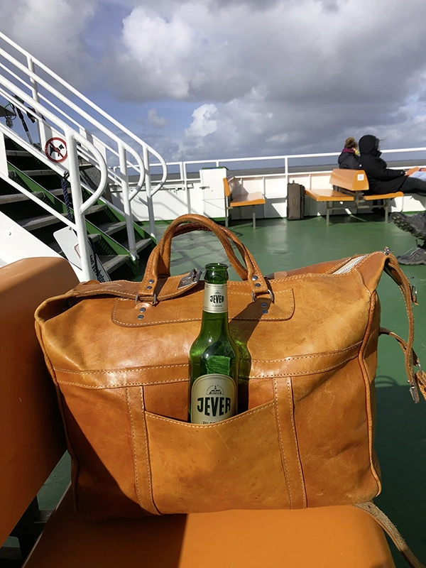 Eine braune Ledertasche steht auf einem Schiff, darin eine grüne Flasche. Im Hintergrund sitzen Personen.