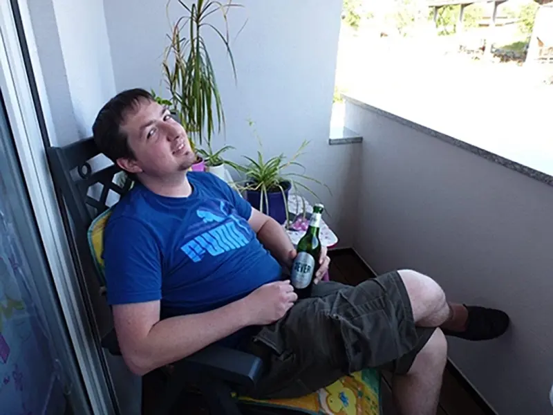 Ein Mann sitzt auf einem Balkonstuhl und hält eine Flasche in der Hand.