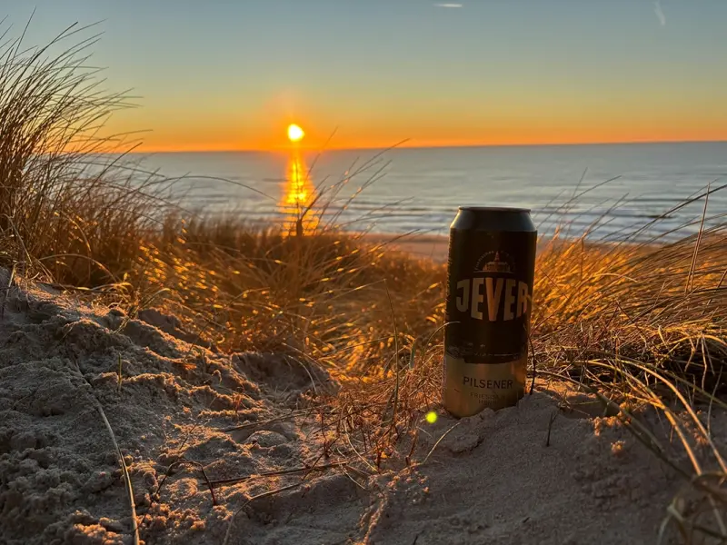 Eine Getränkedose steht im Sand vor einem Sonnenuntergang am Meer. Gräser bilden den Vordergrund.