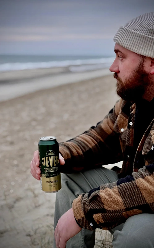 Ein Mann mit Bart und Mütze hält ein Getränk am Strand. Er sitzt seitlich zur Kamera und blickt auf das Meer.