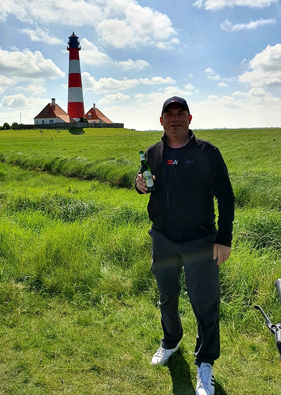Ein Mann steht auf einer Wiese vor einem roten und weißen Leuchtturm und hält eine Flasche.