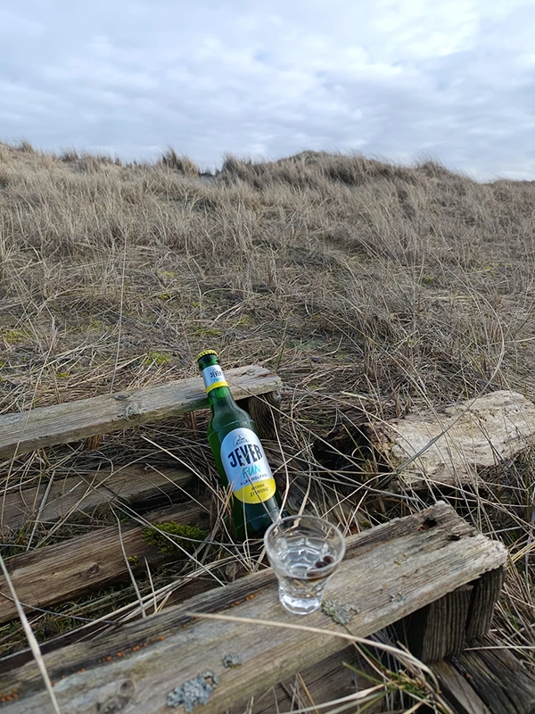 Eine Flasche und ein Glas stehen auf einem alten Holzbalken vor einem Dünengrasfeld.