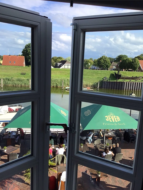 Blick aus einem offenen Fenster auf eine belebte Terrasse an einem Fluss, umgeben von grünen Wiesen und Häusern.