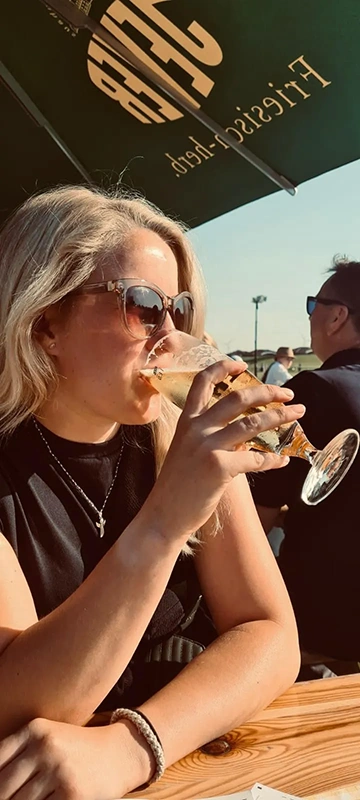 Eine blonde Frau trinkt aus einem Bierglas an einem sonnigen Tag im Freien.