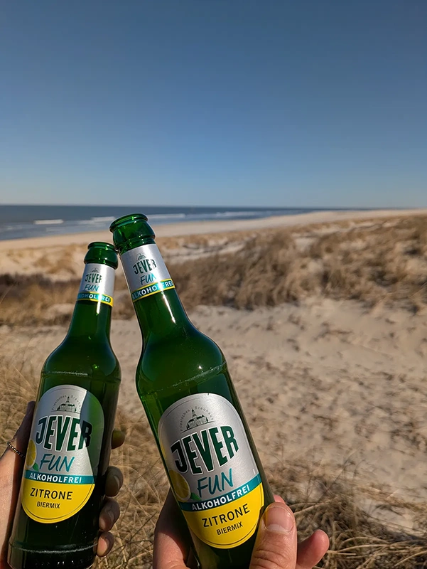 Zwei Hände halten alkoholfreie Getränkeflaschen an einem sonnigen Strand.