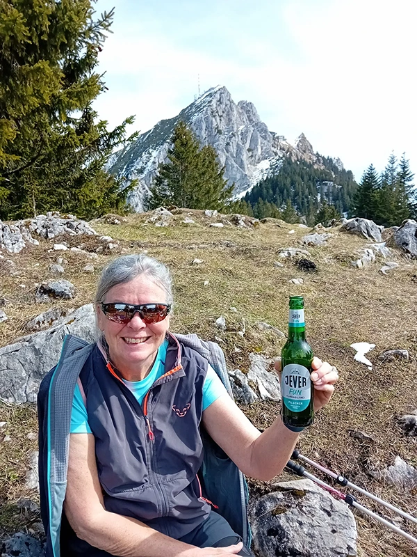 Eine lachende Person sitzt im Freien mit einem Getränk in der Hand vor einem Berg.