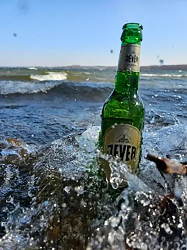 Eine grüne Flasche steht im Wasser am Meer, umgeben von spritzenden Wellen.