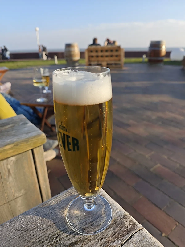 Ein Glas Bier steht auf einem Holztisch, im Hintergrund eine Küstenlandschaft.