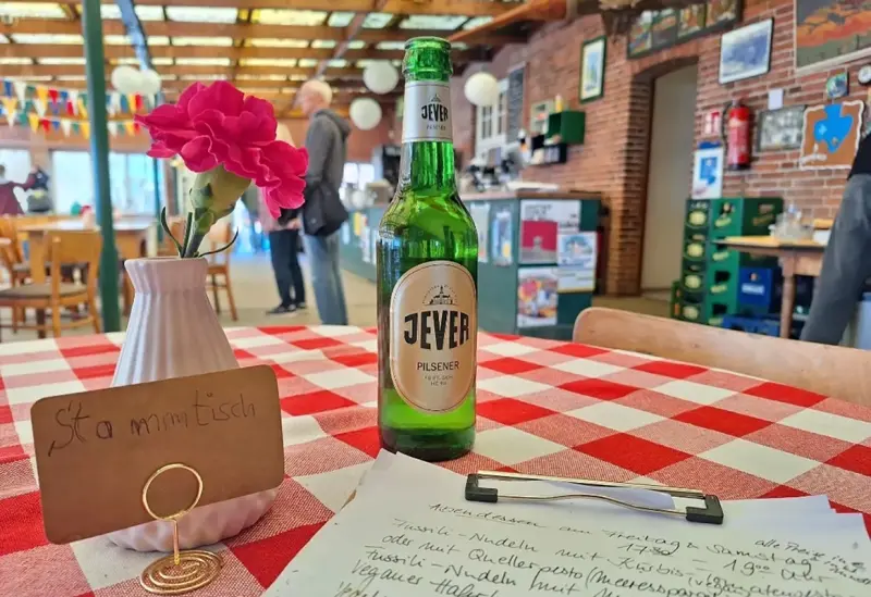 Ein grünes Bierflasche steht auf einem Tisch mit rot-weiß kariertem Tischtuch neben einer rosa Blume.