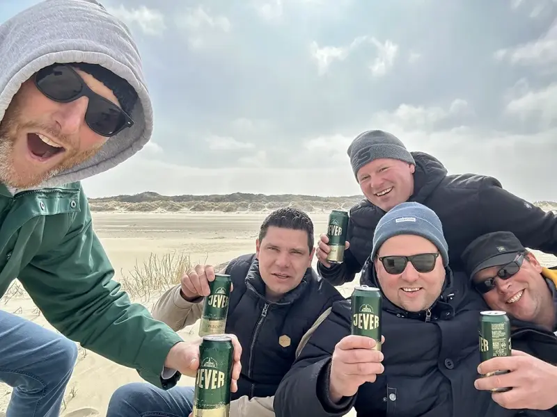 Fünf Männer lachen und halten Getränkedosen am Strand in die Kamera. Sie tragen Winterkleidung.