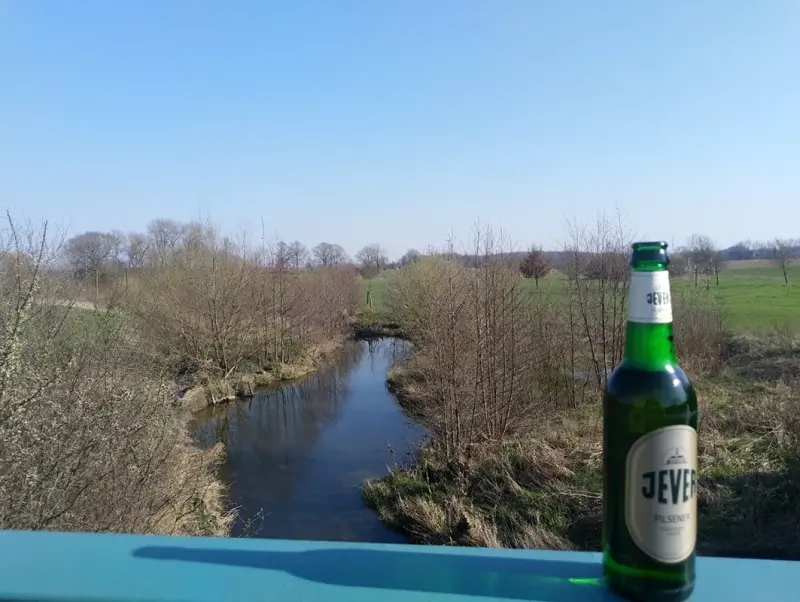 Eine grüne Flasche steht im Vordergrund, dahinter ein ruhiger Fluss in einer Wiesenlandschaft.