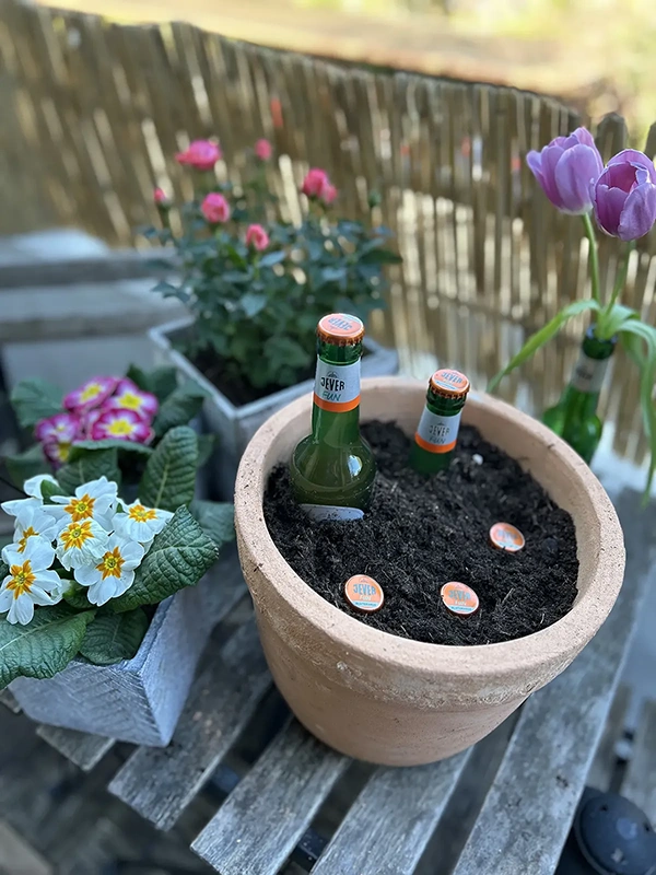 Ein Blumentopf mit Erde und drei kleinen Bierflaschen steht auf einem Holztisch neben blühenden Pflanzen.