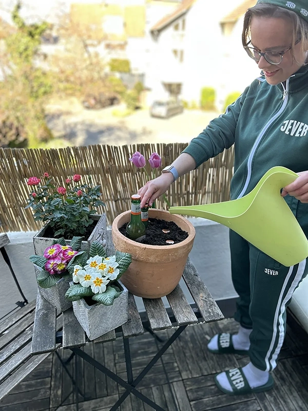 Eine Frau gießt Erde in einen Blumentopf auf einem Balkon. Sie trägt Freizeitkleidung und steht neben blühenden Pflanzen.