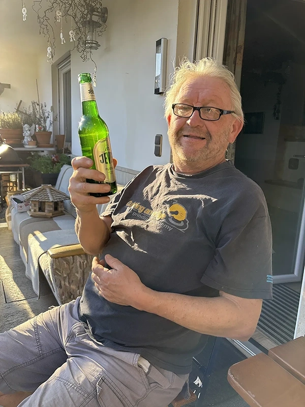 Ein älterer Mann mit Brille sitzt draußen und hält lachend eine grüne Flasche hoch.