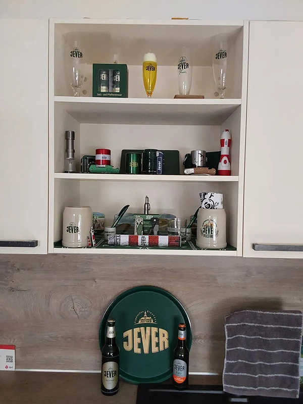 Eine weiße Regalwand präsentiert verschiedene Gläser und Utensilien mit grünen Akzenten; vorne stehen zwei Flaschen und ein rundes Schild.