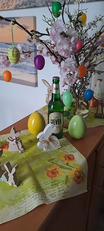 Ein Tisch ist mit einer blumigen Tischdecke bedeckt, darauf stehen bunte Ostereier, Holzhasen und eine grüne Flasche.