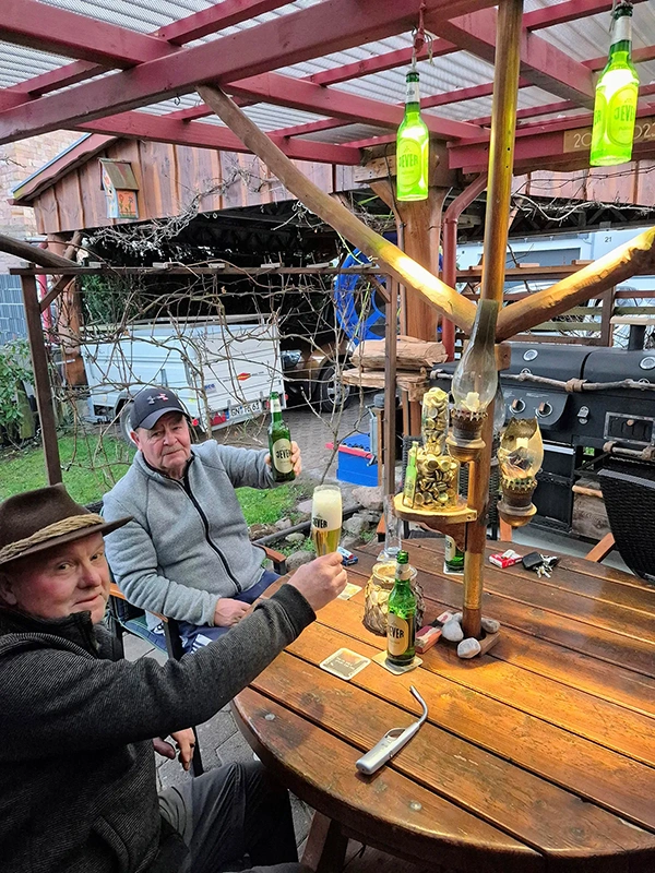 Zwei Männer sitzen an einem rustikalen Holztisch und stoßen mit Getränken an.