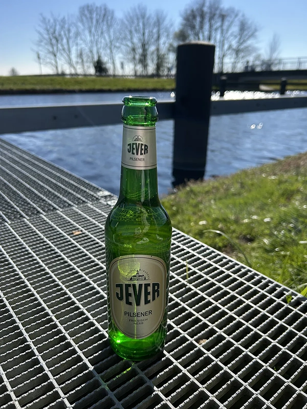 Grüne Bierflasche steht auf einem Gittertisch mit Fluss und Bäumen im Hintergrund.