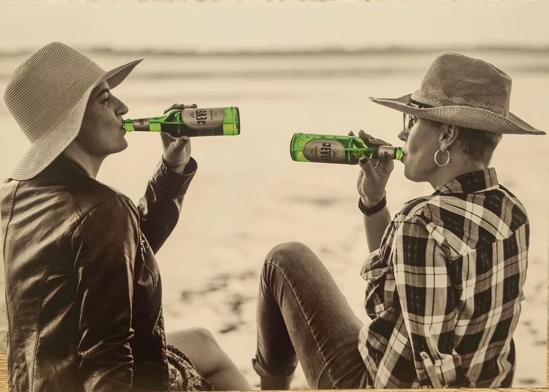 Zwei Personen trinken Getränke aus Flaschen am Strand. Sie tragen Hüte und sitzen sich gegenüber.