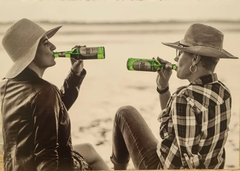 Zwei Personen trinken Getränke aus Flaschen am Strand. Sie tragen Hüte und sitzen sich gegenüber.
