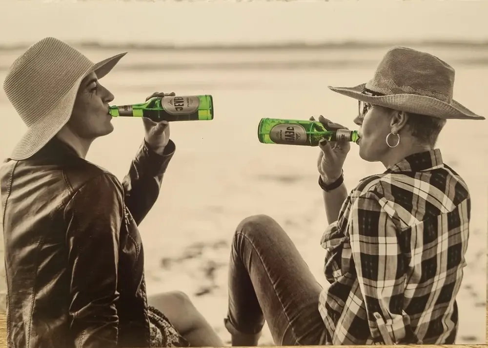 Zwei Personen trinken Getränke aus Flaschen am Strand. Sie tragen Hüte und sitzen sich gegenüber.
