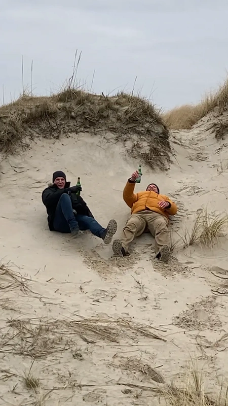 Zwei Personen sitzen lachend mit Getränken in einer Düne. Sie tragen warme Kleidung.