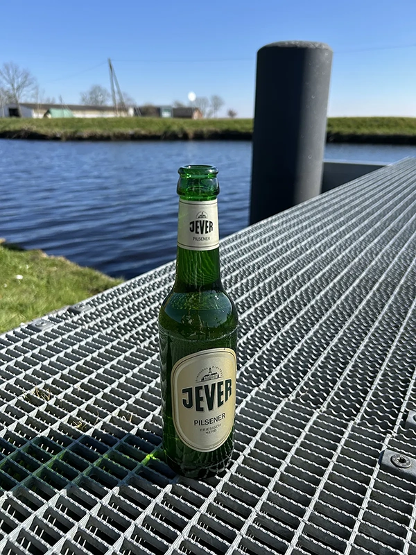 Eine grüne Bierflasche steht auf einem metallischen Gittertisch an einem Fluss.