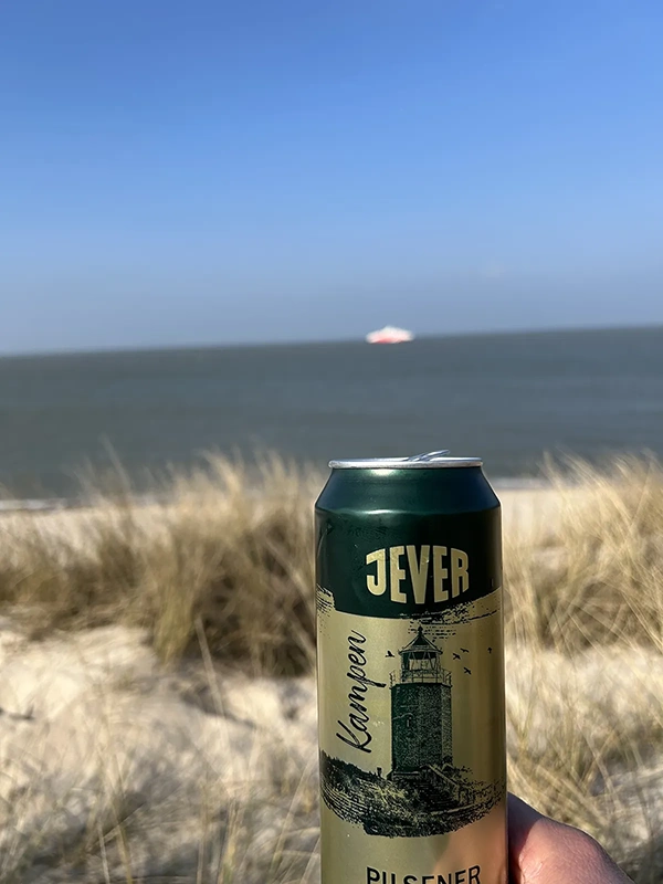 Eine Hand hält eine Getränkedose vor einer Meereskulisse und Dünengras. Im Hintergrund ist ein unscharfes Schiff zu erkennen.