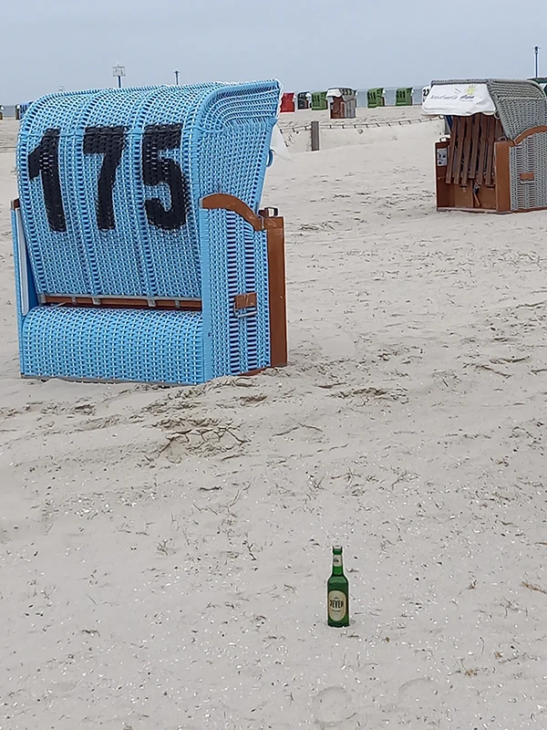Ein blauer Strandkorb steht auf Sand, Nummer 175, neben einer leeren Flasche.