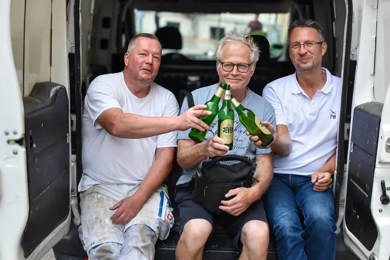 Drei Männer sitzen in einem offenen Transporter und prosten sich mit Flaschen zu. Sie sind lächelnd und tragen lässige Kleidung.