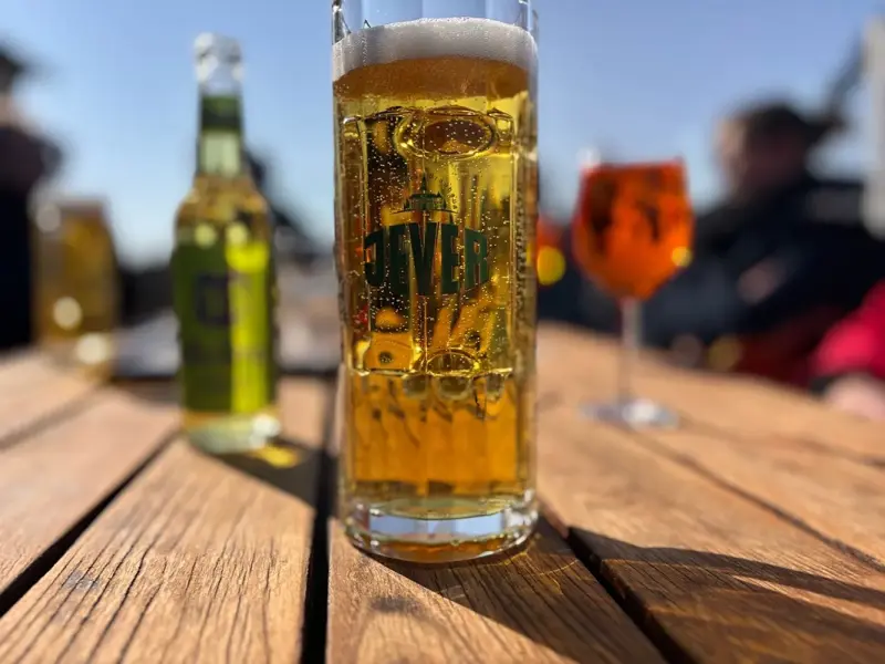 Ein Glas Bier und eine Flasche stehen scharf im Vordergrund auf einem Holztisch.