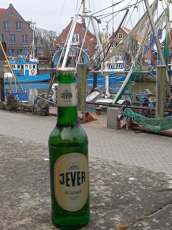 Eine grüne Bierflasche steht im Vordergrund, dahinter liegen Boote an einem Kai.