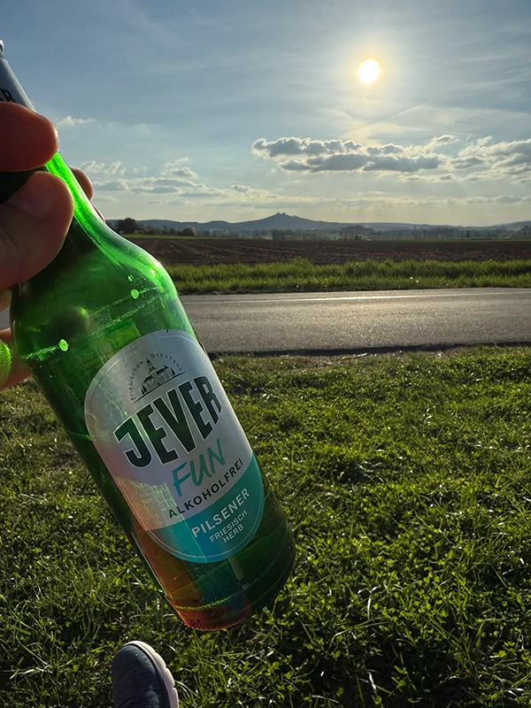 Eine grüne Glasflasche wird vor einer ländlichen Sonnenuntergangsszene gehalten.