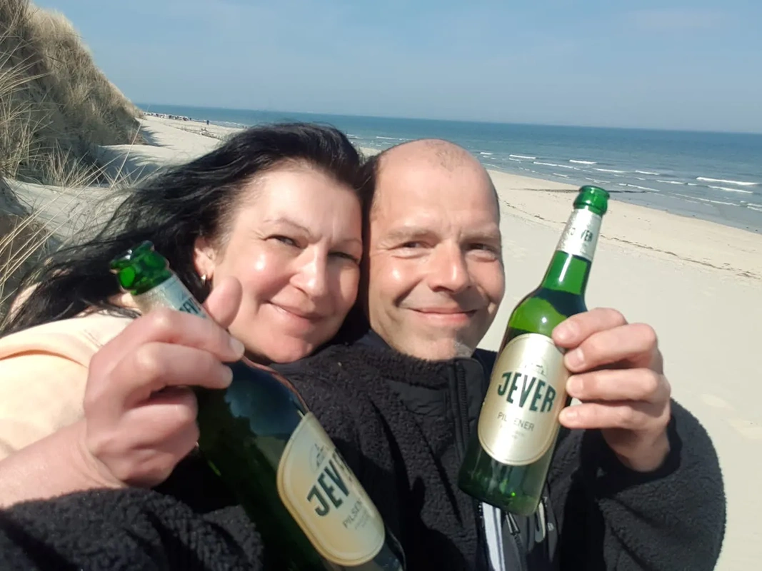 Ein Mann und eine Frau halten lächelnd Bierflaschen an einem sonnigen Strand.