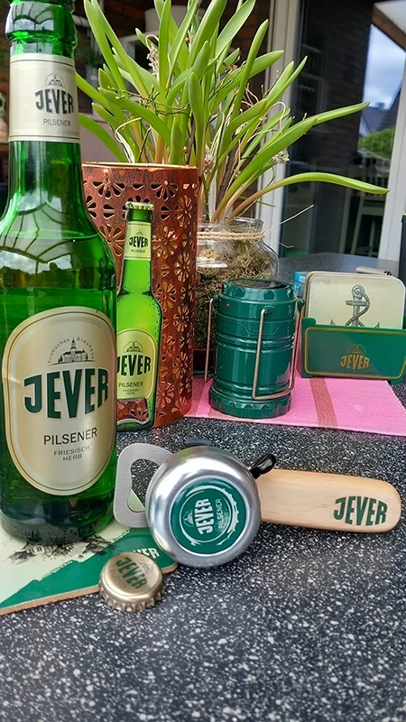 Eine grüne Bierflasche steht neben einer Pflanze auf einem Tisch. Verschiedene Werbeartikel einer Biermarke sind zu sehen.