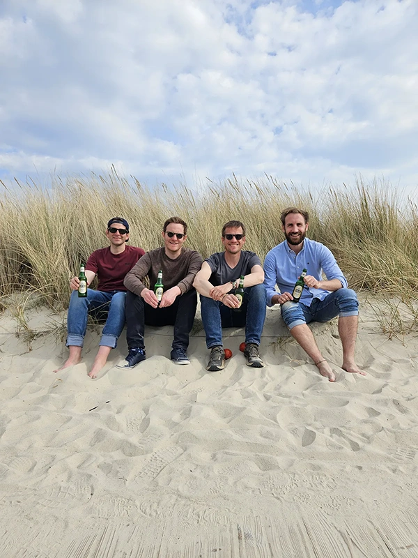 Vier Männer sitzen im Sand mit Gräsern im Hintergrund und halten Getränke.